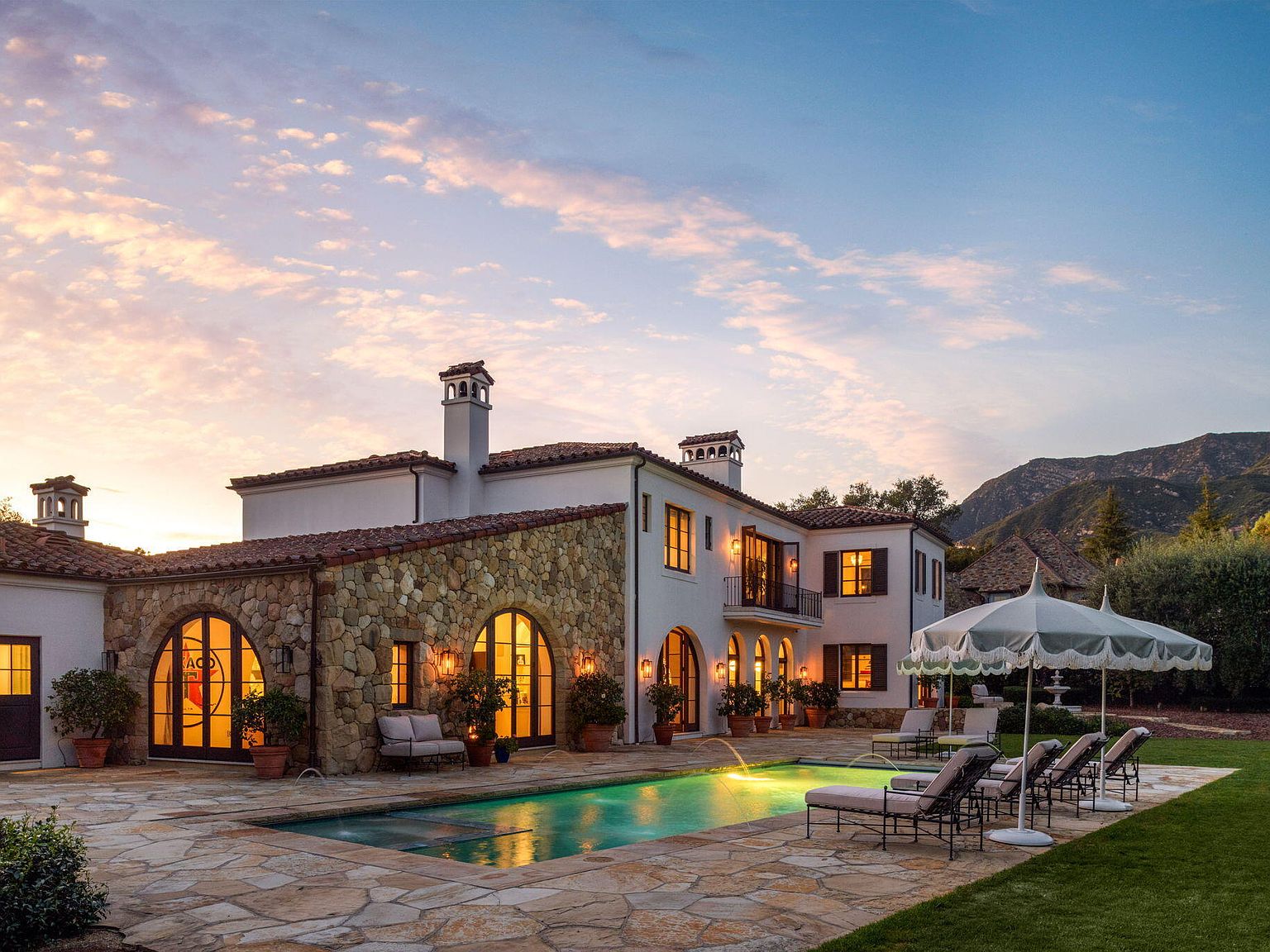 Mediterranean estate at twilight in Montecito with stone walls, lit windows, pool, and the Santa Ynez mountains beyond